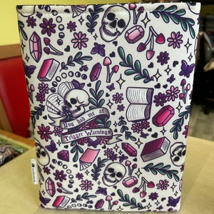 Puede incluir: Una funda de libro blanca con un patrón colorido de calaveras, flores, corazones y libros. El texto "You had me at Trigger Warnings" está impreso en la funda.