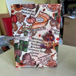 May include: A rectangular fabric item featuring a vibrant autumn design. The design incorporates illustrations of pumpkins, pie, bread, and text including "Pumpkin Pie" and "Po-Ta-Toes". The item is likely a decorative cover or case.