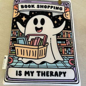 May include: A white ghost with a big smile is holding a shopping basket full of books. The ghost is in front of a bookshelf with colourful books. The text on the image says "Book Shopping is my Therapy".