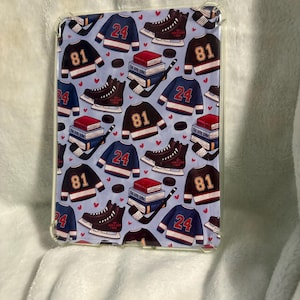May include: A light blue tablet case featuring a repeating pattern of hockey jerseys with the numbers 24 and 81, hockey skates, pucks, hockey sticks, and books. The case has a clear plastic border.
