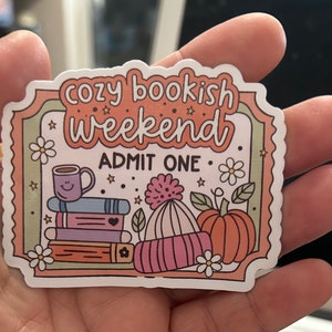 May include: A sticker with a cosy theme, featuring the text "cozy bookish weekend" and "ADMIT ONE." The design includes a stack of books, a mug, a knit hat, and a pumpkin, all in soft pastel colours.