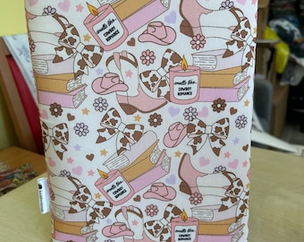 May include: A white book cover with a repeating pattern of pink cowboy boots, brown bows, pink hearts, and stars. The pattern also includes stacks of books and candles with the text "smells like cowboy romance".