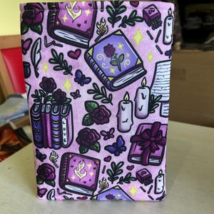 May include: A pink fabric box with a repeating pattern of purple books, roses, butterflies, candles, and leaves. The books have gold accents and the word "LOVE" is visible on the spines. The theme is fantasy.