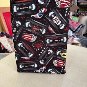 May include: A black book sleeve with a repeating pattern of VHS tapes, featuring horror film titles such as "Halloween" and "Scream." The design incorporates images of knives and blood splatters, creating a spooky aesthetic.