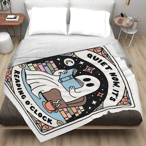 May include: A white blanket featuring a cartoon ghost reading a book, with the text "Reading O'Clock" and "Quiet Now, It's". The design includes a coffee cup, books, stars, and a crescent moon. The blanket is placed on a bed.