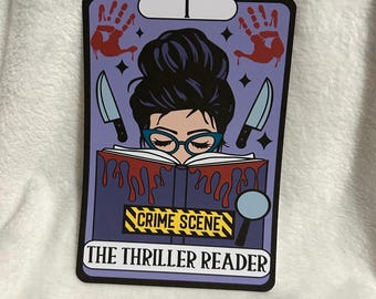 Puede incluir: Una carta de tarot morada con una mujer leyendo un libro. El libro está etiquetado como "Escena del crimen" y la carta dice "El lector de thrillers". La mujer tiene el pelo oscuro en un moño y lleva gafas. Hay huellas de manos ensangrentadas y cuchillos en la carta.