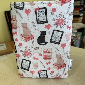 May include: A white book sleeve with a repeating pattern of cats, books, hearts, and floral elements in pink and red. The sleeve features the word "LOVE" on a tablet and a coffee cup design.