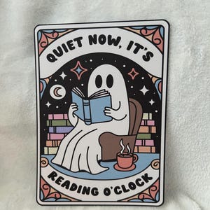 Pode incluir: Adesivo retangular com um fantasma de desenho animado lendo um livro. O adesivo tem o texto "QUIET NOW, IT'S READING O'CLOCK" e uma borda colorida com estrelas, uma lua e uma xícara de café.