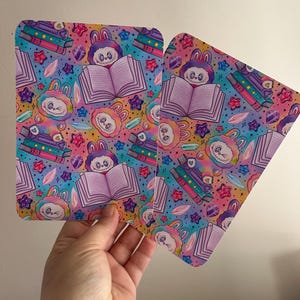 Puede incluir: Dos objetos rectangulares con un patrón colorido que presenta personajes de dibujos animados, libros y cristales. El fondo tiene un degradado de arcoíris. Un objeto está parcialmente abierto. Los objetos son sostenidos por una mano.