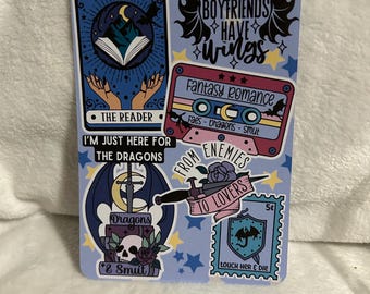 Op de afbeelding: Een set van zes stickers met fantasy- en romantische thema's. De stickers zijn gedrukt op een witte achtergrond met blauwe, roze en zwarte accenten. De stickers bevatten illustraties van draken, een boek, een cassettebandje en een postzegel. De tekst op de stickers is "My book boyfriends have wings", "Fantasy Romance", "Faes - Dragons - Smut", "From Enemies to Lovers", "Dragons & Smut", en "Touch Her & Die".