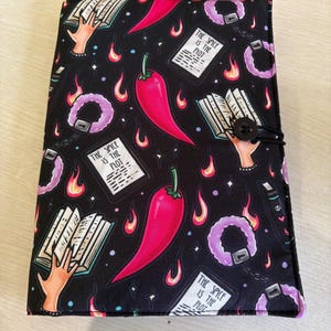 May include: A black fabric book cover featuring a repeating pattern of red chilli peppers, open books with hands, purple fuzzy handcuffs, and white rectangular cards with the text "THE SPICE IS THE PLOT". The cover includes a black button and elastic closure.