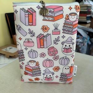 Puede incluir: Una bolsa blanca con cremallera con temática de Halloween. La bolsa presenta un patrón repetitivo de libros, calabazas, fantasmas y hojas de otoño. El tirador de la cremallera es un pequeño amuleto de calabaza.