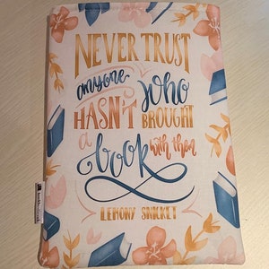 Puede incluir: Una funda de libro blanca con un estampado floral en melocotón, rosa y azul. El texto dice: "Nunca confíes en nadie que no haya traído un libro consigo" de Lemony Snicket. Se ve una pequeña etiqueta con texto.