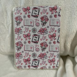 Könnte beinhalten: Eine transparente Schutzhülle für einen E-Reader mit einem rosa-weißen Muster. Das Design umfasst Schleifen, Rosen, offene Bücher mit Herzen, Gänseblümchen und den Text "Romance Book Club".
