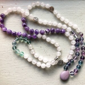 May include: A collection of beaded necklaces and a bracelet. The necklaces feature white, purple, and green beads, with silver-colored accents. One necklace has a teardrop-shaped pendant. The jewelry is arranged on a white surface.