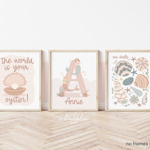 May include: Three printable wall art prints in a pink and blue colour scheme. The first print features a pearl inside an open seashell with the text "the world is your oyster!" The second print features a mermaid swimming around the letter "A" with the name "Annie" below. The third print features a variety of seashells in different colours and patterns with the text "sea shells".