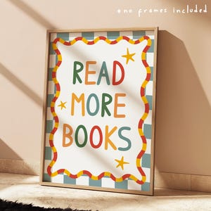 May include: A framed print with the words "READ MORE BOOKS" in colourful, playful lettering. The print has a white background with a wavy red and yellow border, and a blue and white chequered pattern. The frame is a light wood colour.