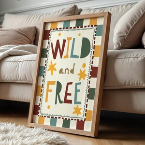 May include: A framed art print with the words "WILD and FREE" in colourful, playful lettering. The artwork features a cream-coloured background with a decorative border and a wooden frame. The print is leaning against a beige sofa.