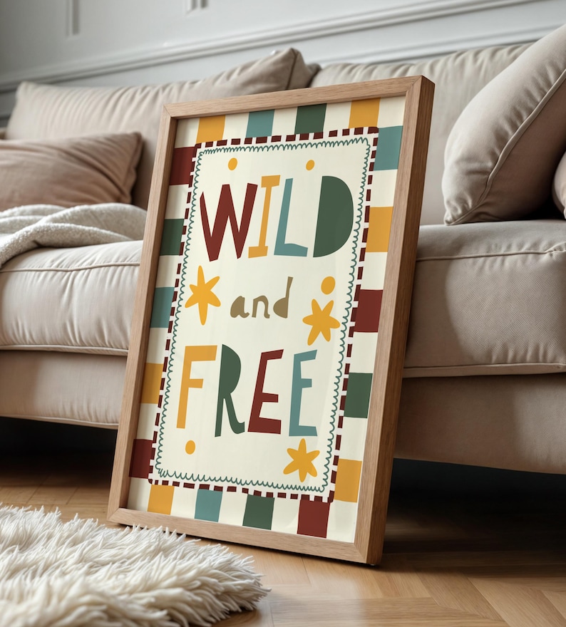 May include: A framed art print with the words "WILD and FREE" in colourful, playful lettering. The artwork features a cream-coloured background with a decorative border and a wooden frame. The print is leaning against a beige sofa.