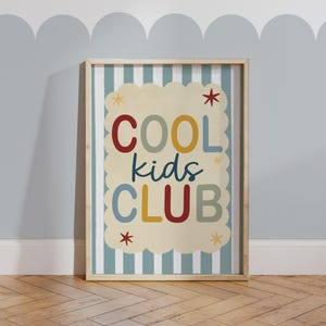 May include: A framed print with the words "COOL kids CLUB" in colorful letters. The background of the print is a cream color with a scalloped edge, set against a striped blue and white background. The frame is a light wood color.