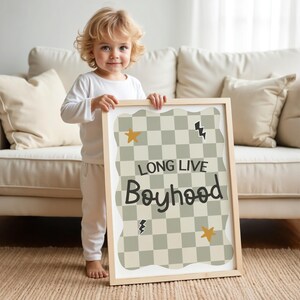 May include: A child is holding a framed print with a chequered background and the text "Long Live Boyhood". The print has a wavy border and features yellow stars and lightning bolts.