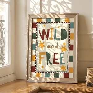 May include: A framed art print displaying the words "WILD and FREE" in a vibrant, multi-coloured design. The artwork has a cream background with a checkered border in red, yellow, green, and blue. The frame is a light wood colour.