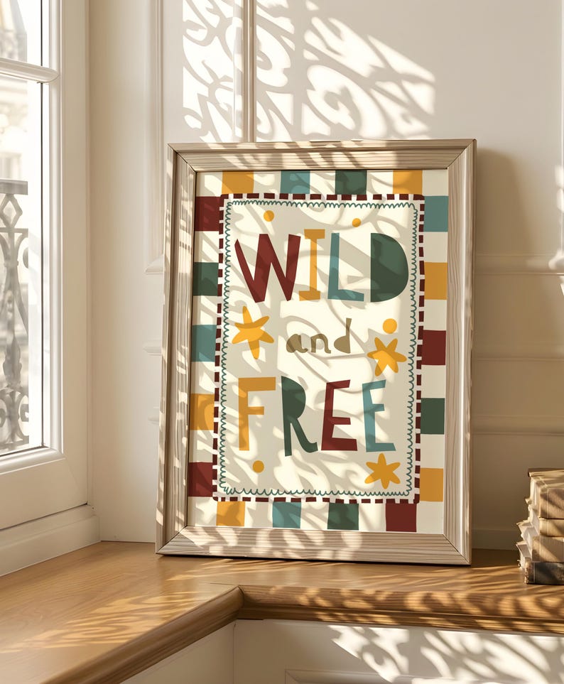 May include: A framed art print displaying the words "WILD and FREE" in a vibrant, multi-coloured design. The artwork has a cream background with a checkered border in red, yellow, green, and blue. The frame is a light wood colour.
