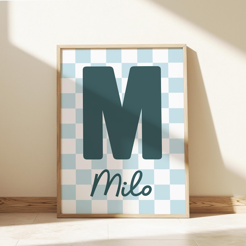 Puede incluir: Un estampado enmarcado con una gran letra "M" de color verde azulado sobre un fondo a cuadros azul claro y blanco. El nombre "Milo" est&aacute; escrito en cursiva debajo. El marco es de color madera clara.