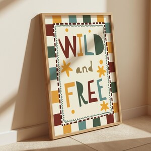 May include: A framed art print with the words "WILD and FREE" in colourful, playful lettering. The artwork features a cream-coloured background with a checkered pattern in shades of brown, green, blue, and yellow. The frame is made of light-coloured wood.