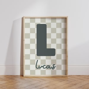 Puede incluir: Un estampado enmarcado con una gran letra "L" gris oscuro sobre un fondo a cuadros de cuadrados grises claros y crema. El nombre "Lucas" est&aacute; escrito en cursiva debajo. El marco es de color madera clara.