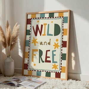 May include: A framed print with the words "WILD and FREE" in colourful letters. The artwork has a cream background with a chequered border in various colours. The frame is made of light-coloured wood. A vase with dried plants and an open magazine are in the foreground.
