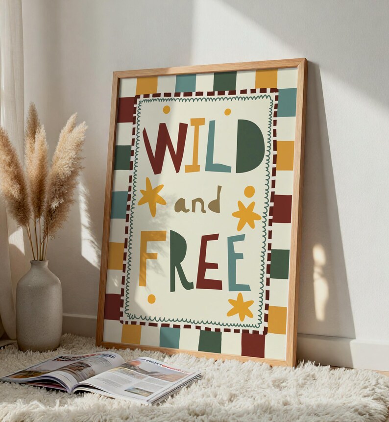 May include: A framed print with the words "WILD and FREE" in colourful letters. The artwork has a cream background with a chequered border in various colours. The frame is made of light-coloured wood. A vase with dried plants and an open magazine are in the foreground.