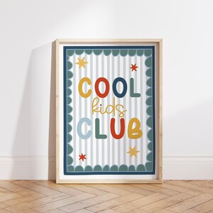 May include: A framed print with the words "COOL kids CLUB" in colorful, playful lettering. The design features a striped background, star accents, and a scalloped border, all within a light wood frame.
