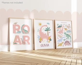 May include: Three framed prints with a pink dinosaur theme. The first print says "ROAR" in pink letters with dinosaurs around the letters. The second print has a pink dinosaur with the name "Olivia" below it. The third print says "Dinosaurs" with a variety of dinosaurs in different colors.