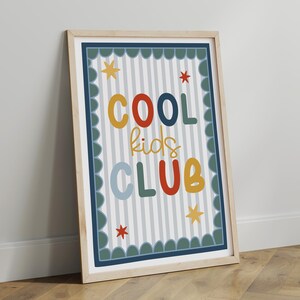 May include: A framed print with the words "COOL kids CLUB" in colorful, playful lettering. The background has vertical stripes, and the frame is light wood. Decorative stars and a scalloped border complete the design.