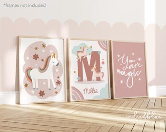 May include: Three pink framed prints with a unicorn theme. The first print features a white unicorn with a rainbow mane and tail on a pink background with gold stars. The second print features a large pink letter M with a unicorn on top and a rainbow at the bottom. The third print features the text "You are magic" in white on a pink background.