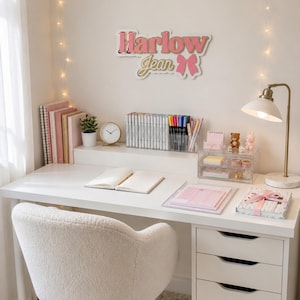 Pu&ograve; includere: Una scrivania bianca con una sedia in sherpa bianca, una lampada dorata e un cartello con la scritta "Harlow Jean". La scrivania ha una pila di quaderni rosa, una piccola pianta, un orologio e un organizer trasparente con pennarelli e penne. C'&egrave; un piccolo tappeto sotto la sedia.