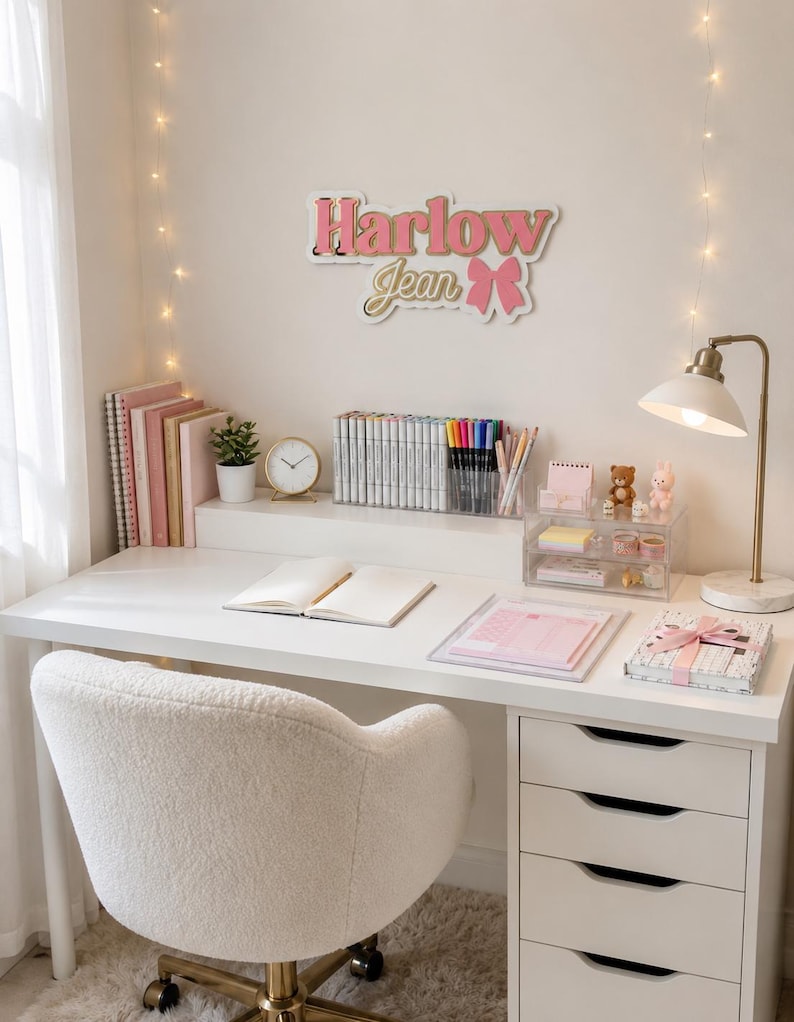 Pu&ograve; includere: Una scrivania bianca con una sedia in sherpa bianca, una lampada dorata e un cartello con la scritta "Harlow Jean". La scrivania ha una pila di quaderni rosa, una piccola pianta, un orologio e un organizer trasparente con pennarelli e penne. C'&egrave; un piccolo tappeto sotto la sedia.