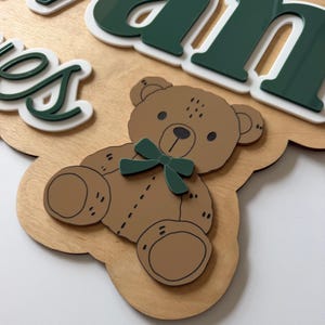 Puede incluir: Letrero de madera con un oso de peluche marr&oacute;n claro con una pajarita verde. El letrero tiene el nombre "James" en letras verdes y blancas. El letrero es de madera y tiene un color marr&oacute;n claro.
