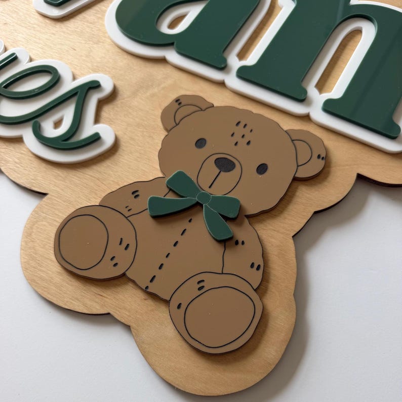 Puede incluir: Letrero de madera con un oso de peluche marr&oacute;n claro con una pajarita verde. El letrero tiene el nombre "James" en letras verdes y blancas. El letrero es de madera y tiene un color marr&oacute;n claro.