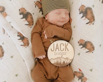 May include: A newborn baby wearing a brown onesie and a striped hat is lying on a white blanket with a brown wooden sign that says "JACK bridger" with a mountain scene.