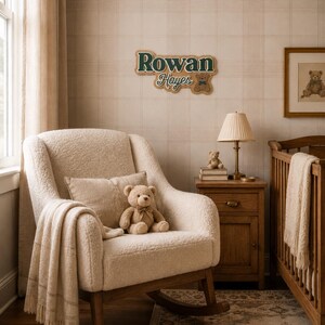 Puede incluir: Un cuarto de beb&eacute; con una mecedora color crema, una cuna de madera y una mesita de noche. Un osito de peluche est&aacute; sentado en la silla, y un letrero en la pared dice "Rowan Hayes". La habitaci&oacute;n tiene una paleta de colores suaves y neutros con una alfombra en el suelo.