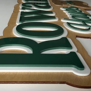 Puede incluir: Letrero de madera con la palabra "Rowan" en letras verdes y blancas superpuestas. El letrero tiene una base de madera marr&oacute;n claro y un borde blanco, creando un efecto 3D. Las letras est&aacute;n en una fuente moderna y en negrita.