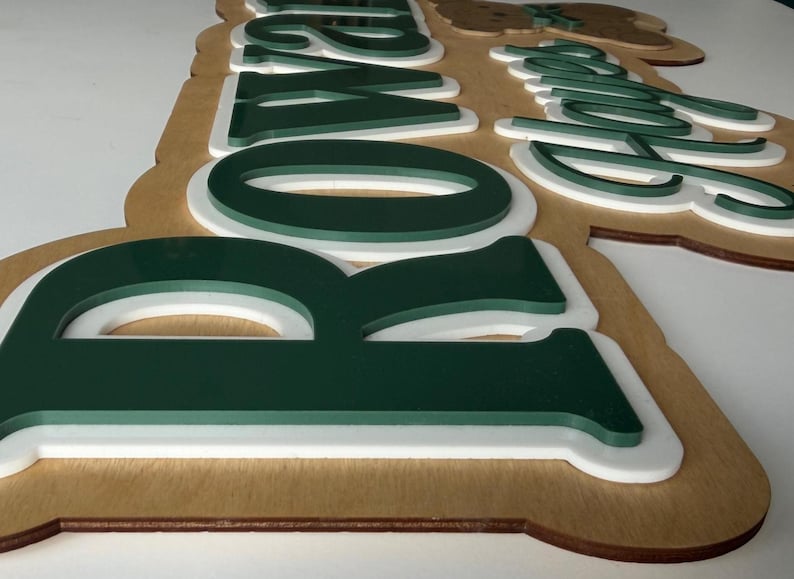 Puede incluir: Letrero de madera con la palabra "Rowan" en letras verdes y blancas superpuestas. El letrero tiene una base de madera marr&oacute;n claro y un borde blanco, creando un efecto 3D. Las letras est&aacute;n en una fuente moderna y en negrita.