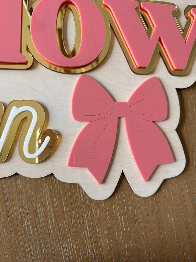 Pu&ograve; includere: Un cartello in legno con la scritta "BOW" in lettere rosa con bordi dorati. Un fiocco rosa &egrave; posizionato sotto le lettere. Il cartello ha un bordo smerlato ed &egrave; su una superficie di legno.