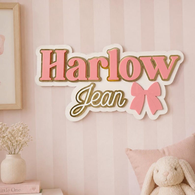 Pu&ograve; includere: Un'insegna con nome rosa e oro con la scritta "Harlow Jean" e un fiocco. L'insegna &egrave; montata su una parete a righe rosa e bianche. Le lettere sono rosa con un contorno dorato, un oggetto decorativo.