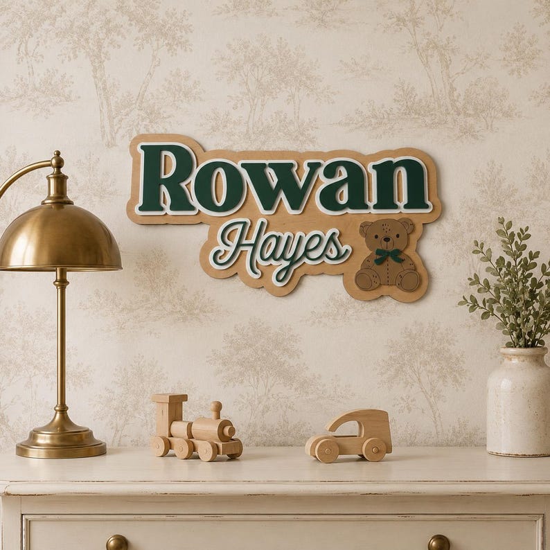 Puede incluir: Un letrero de madera con el nombre "Rowan Hayes" en letras verdes y blancas, con una ilustraci&oacute;n de un oso de peluche. El letrero se muestra encima de una c&oacute;moda blanca con una l&aacute;mpara de lat&oacute;n, un tren y un coche de madera.