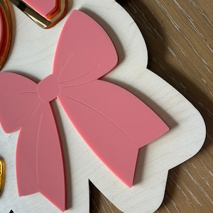 Pu&ograve; includere: Un fiocco rosa chiaro &egrave; il punto focale di questo pezzo decorativo in legno. Il fiocco &egrave; sovrapposto a una base di legno bianca con bordi smerlati. Accenti color oro sono visibili sul lato sinistro dell'immagine. Lo sfondo &egrave; una superficie in legno.