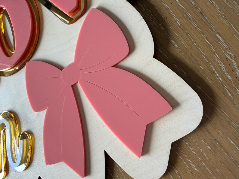 Pu&ograve; includere: Un fiocco rosa chiaro &egrave; il punto focale di questo pezzo decorativo in legno. Il fiocco &egrave; sovrapposto a una base di legno bianca con bordi smerlati. Accenti color oro sono visibili sul lato sinistro dell'immagine. Lo sfondo &egrave; una superficie in legno.