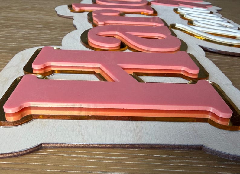 Pu&ograve; includere: Cartello in legno rosa a strati con accenti dorati e arancioni. Il cartello presenta la parola "Bride" in un design a strati. Il cartello &egrave; su una superficie di legno chiaro.